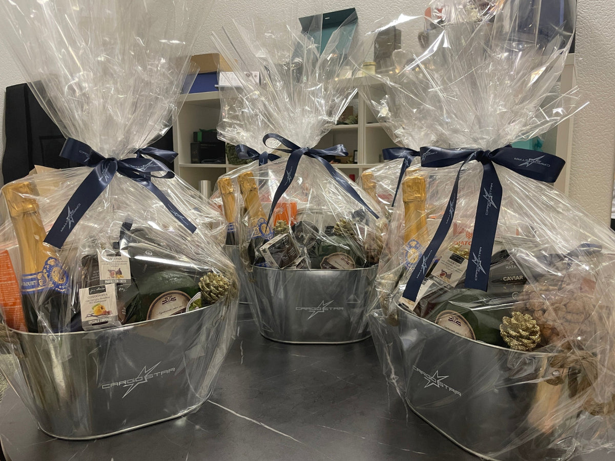 Three gourmet gift baskets in silver metal buckets, wrapped in clear cellophane and tied with navy blue ribbons, displayed on a dark countertop.