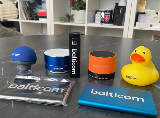 A collection of Balticom-branded smart home and bath accessories arranged on a dark gray marble surface next to a white shelving unit. The items include a yellow rubber duck with an orange beak, a blue rectangular device, an orange cylindrical speaker, a black box, a blue-and-white cylindrical container, a blue shower head, and a clear case with a Balticom logo.