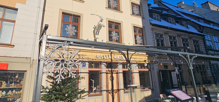 A sunny outdoor street scene featuring historic beige and gray buildings with snow-covered roofs, storefront windows, and a decorative metal snowflake archway in the foreground.