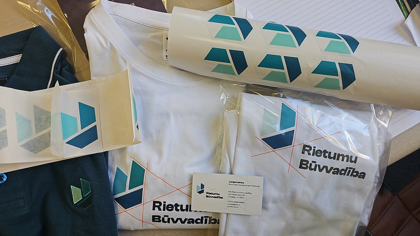 A white athletic t-shirt with a blue and green geometric logo and the text 'Rietumu Bövvadöiba' printed on the front, shown alongside matching printed sleeves, a rolled-up sleeve, and a small brand card.