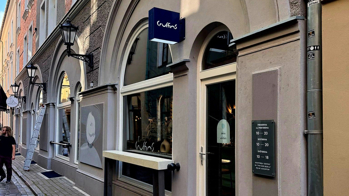 A street view of a storefront with arched windows and door, a blue sign reading 'Café', a cobblestone sidewalk, and a person walking on the left side of the street.